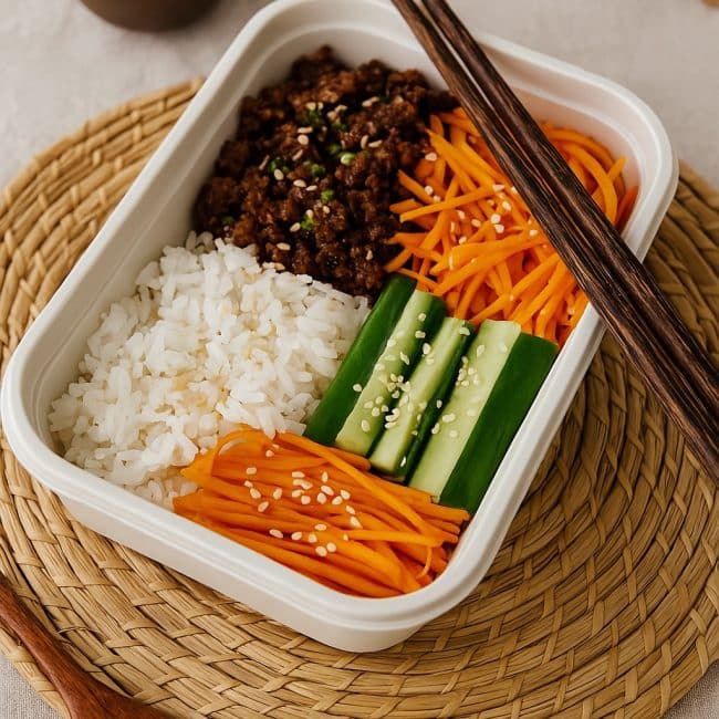 Koreanische Reis Bowl mit Rindfleisch, Gemüse und Sesam, angerichtet in einer stylischen Meal-Prep-Dose mit Holzstäbchen und asiatischer Tischdeko.