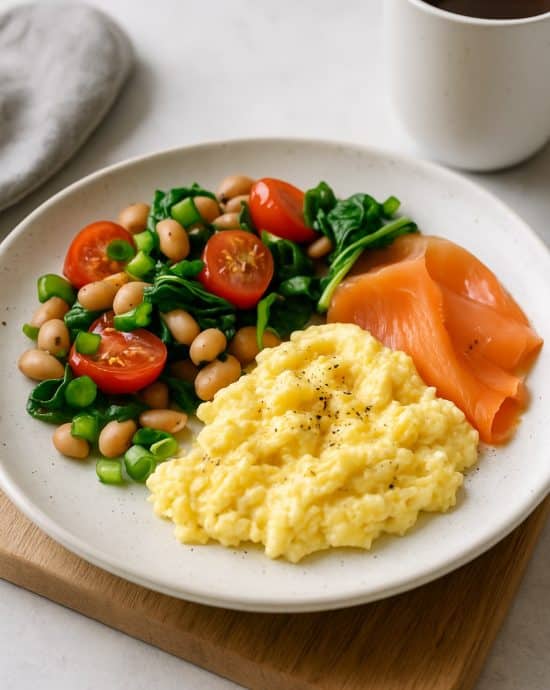 Modern interpretiertes britisches Frühstück mit Rührei, Räucherlachs, weißen Bohnen, Tomaten und Spinat auf einem Frühstücksteller.
