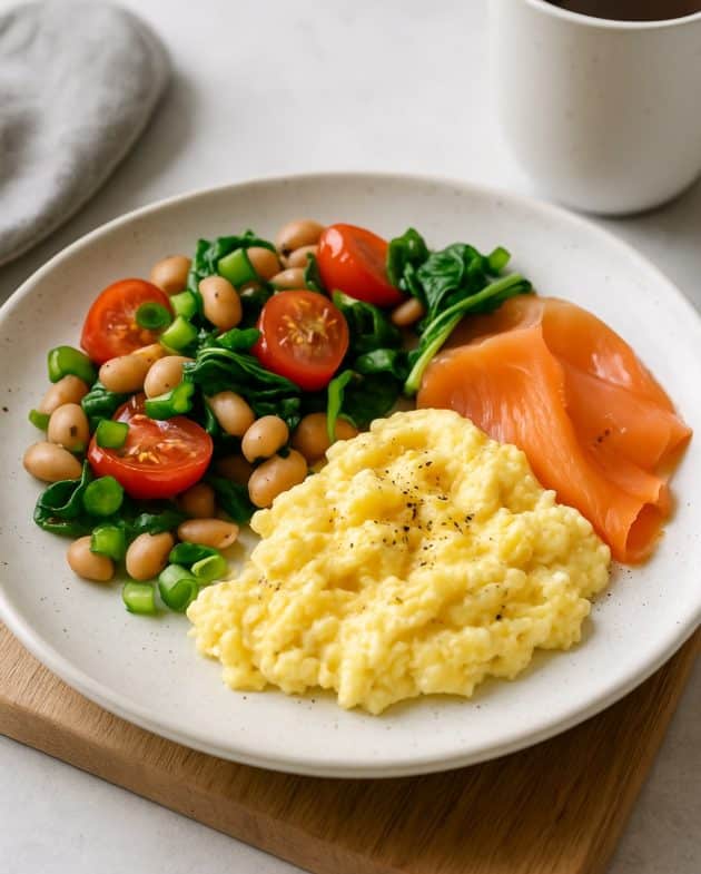 Modern interpretiertes britisches Frühstück mit Rührei, Räucherlachs, weißen Bohnen, Tomaten und Spinat auf einem Frühstücksteller.