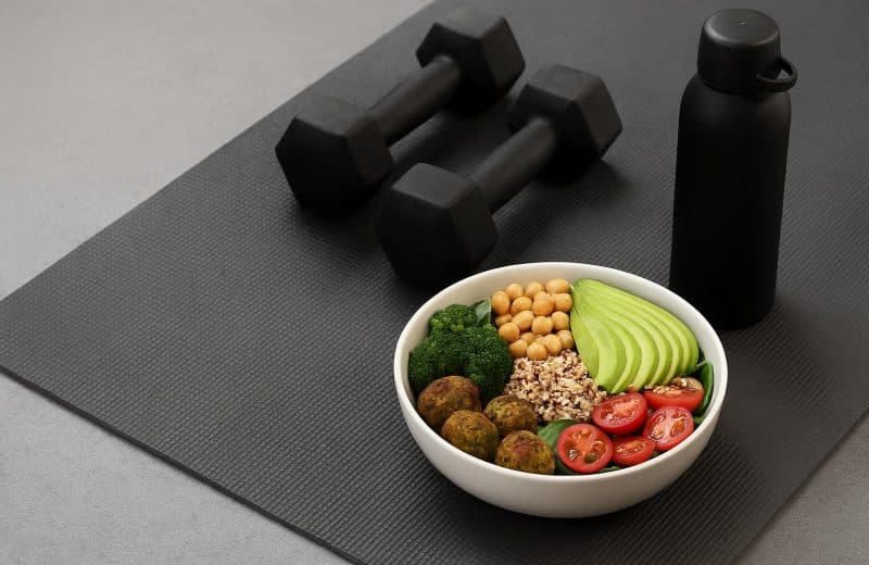 vegane Fitness-Ernährung Bowl mit Gemüse und Tofu auf Fitnessmatte mit Hanteln und Wasserflasche