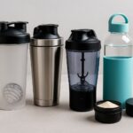 Verschiedene Fitness-Shaker und Trinkflaschen aus Edelstahl, Kunststoff und Glas auf einem Tisch mit Sportzubehör